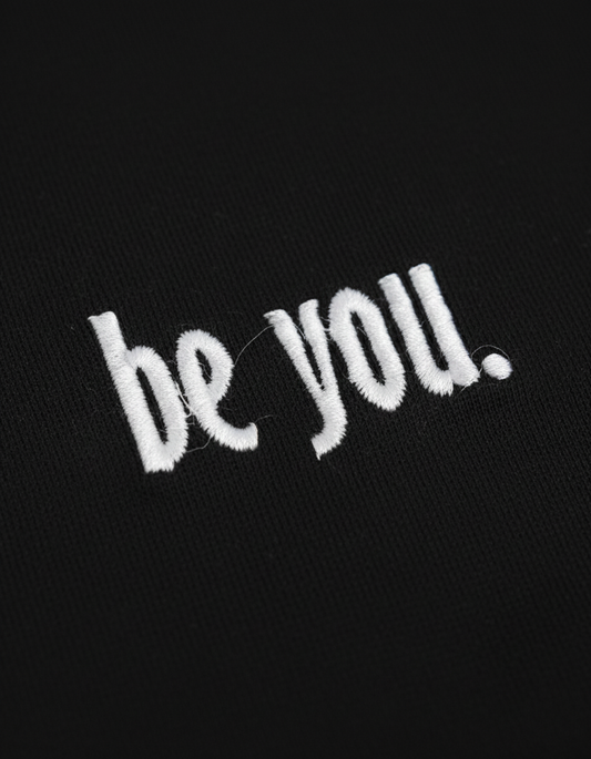Black fabric with white 'be you.' embroidery on a black background