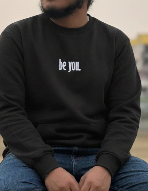 Person wearing a black sweatshirt with 'be you.' text, sitting outdoors.